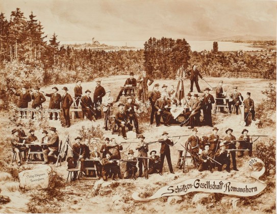 Fotografie: Gruppenporträt der Schützengesellschaft Romanshorn in Landschaft am Bodensee, mit Schloss Romanshorn und der Textilfabrik am See im Hintergrund
