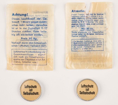 Ansteckknöpfe mit fluoreszierender Beschichtung und der Aufschrift «Luftschutz ist Selbstschutz», in originaler Verpackung mit gedruckter Gebrauchsanleitung